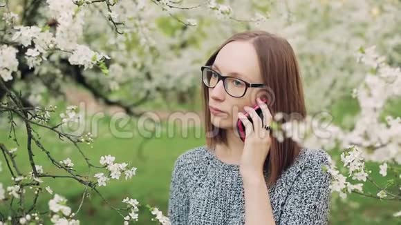 戴眼镜的悲伤女孩在盛开的花园里用手机说话视频的预览图