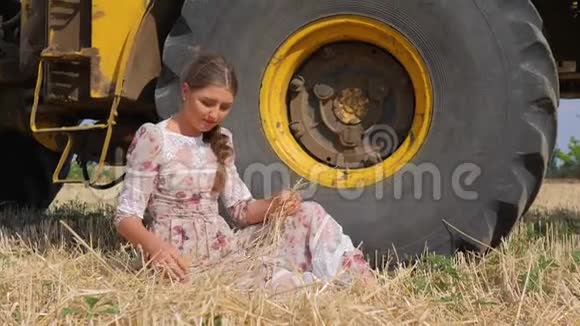 穿着漂亮的年轻女子坐在农村农场的联合收割机的大轮子旁视频的预览图