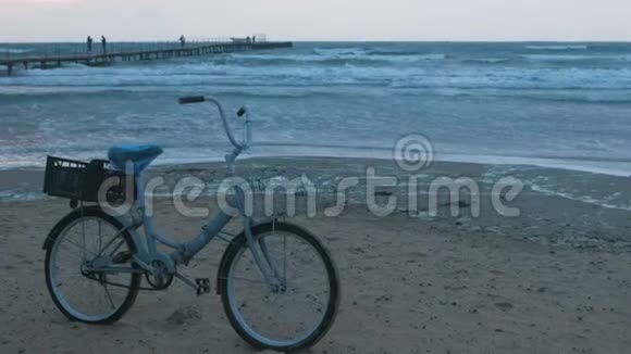 夕阳西下沙滩上的旧自行车带着风暴的大海和泡沫的波浪背景码头上有步行的人视频的预览图
