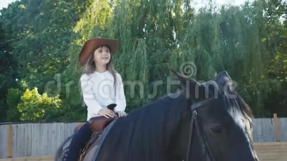 戴帽子的漂亮小女孩骑着一匹黑色的母马在竞技场上4K视频的预览图