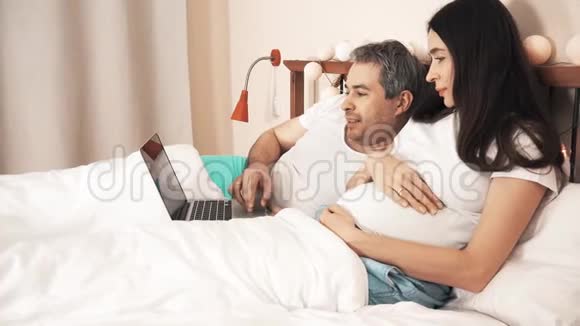 男人和他美丽的怀孕妻子在床上用笔记本电脑视频的预览图