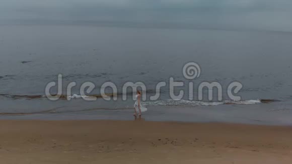 高空俯视图美丽的金发女郎在海边漫步一位穿着白色连衣裙海浪拍打着海面的仙女视频的预览图
