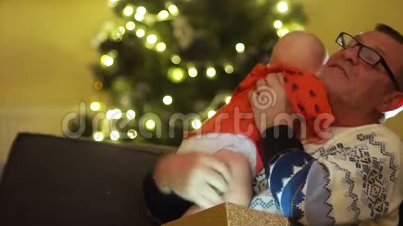 成熟的男人在圣诞树灯的背景下慈爱地拥抱坐在沙发上的孩子视频的预览图