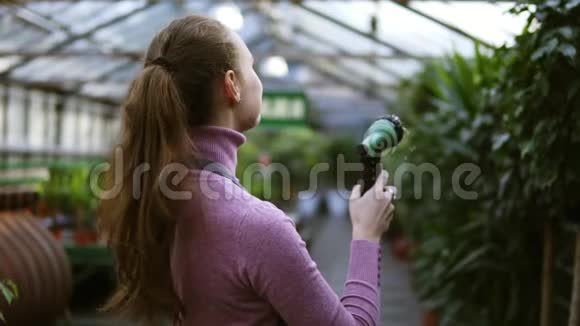 年轻微笑的女园丁穿着统一的浇水植物在温室里用花园软管浇水慢镜头视频的预览图
