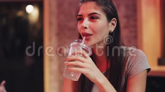 年轻的美女喝着鸡尾酒坐在靠近霓虹灯标志的酒吧或餐馆里视频的预览图