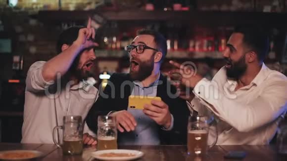 三个老朋友在智能手机上看视频在运动酒吧喝啤酒朋友们笑着讨论视频的预览图