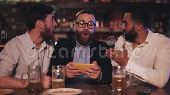 三个老朋友在智能手机上看视频在运动酒吧喝啤酒朋友们笑着讨论视频的预览图