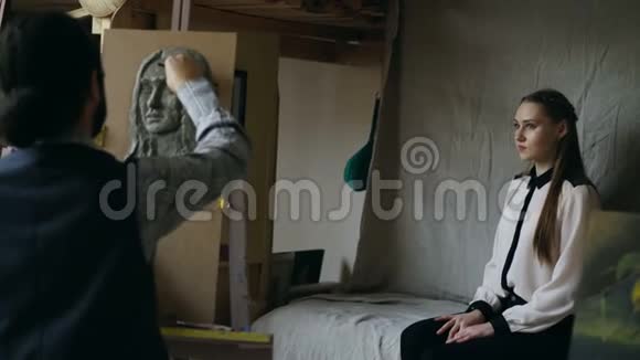 雕塑家在画室里给摆姿势的年轻女子在画布上创作人的面部雕塑视频的预览图