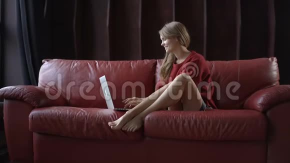 一位漂亮的年轻女士坐在家里的沙发上用手提电脑微笑着看着相机视频的预览图