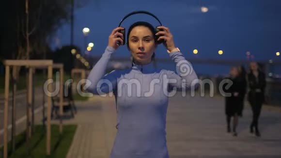 女孩戴上耳机晚上开始穿过城市月光下的运动员训练视频的预览图