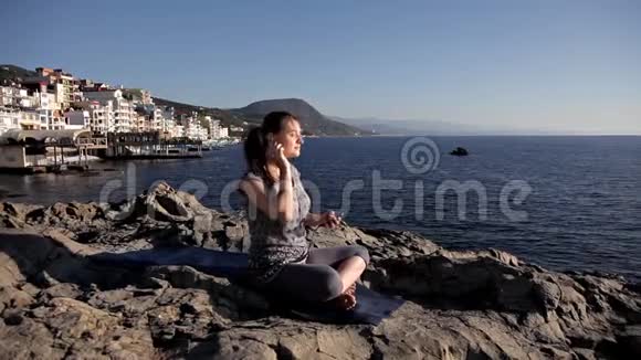 穿着运动服和耳机的女人坐在海上岩石上的莲花位置冥想听音乐瑜伽视频的预览图