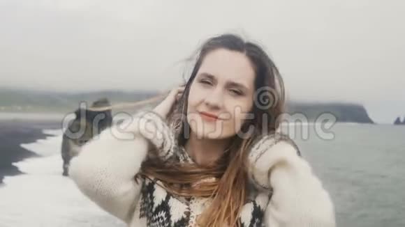 年轻快乐的女人站在黑色海滩上的肖像看着相机微笑着靠近冰岛的巨魔脚趾岩石视频的预览图