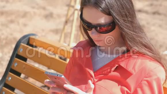 年轻的黑发女孩戴着太阳镜在坐在长凳上的公园里看着手机上的视频视频的预览图
