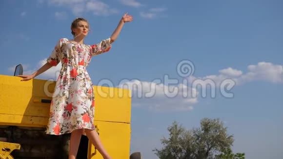 农村女孩穿上衣服在大自然的天空背景下手在拖拉机的帽子上挥舞视频的预览图