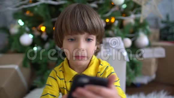 可爱的非常可爱的男孩坐在圣诞树的背景上玩智能手机慢动作儿童视频的预览图