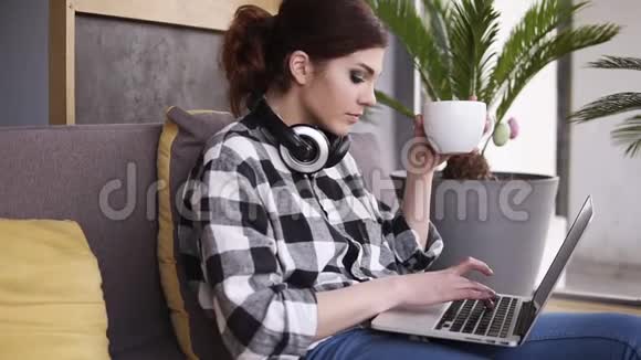 放松迷人的女孩坐在房间的沙发上打字在笔记本电脑上手里拿着一个白色的大杯子女孩喝酒视频的预览图