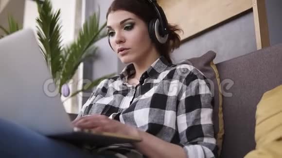 从下面拍摄一个坐在沙发上的迷人的黑发女人戴着耳机的女孩在笔记本电脑上打字微笑着视频的预览图