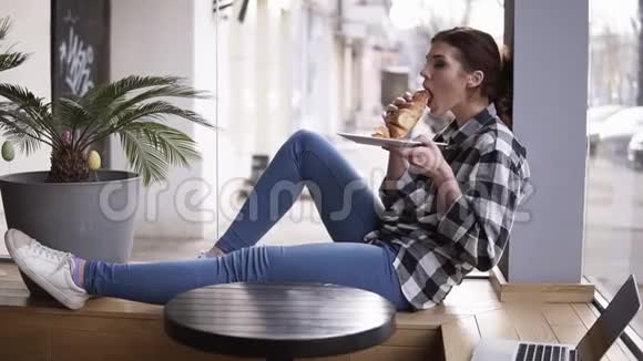 一个年轻漂亮的女孩坐在明亮窗户旁边的木座上的咖啡馆里的侧面景色女孩正在吃视频的预览图