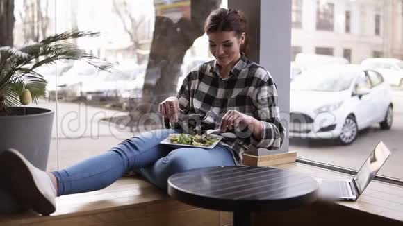 女孩靠在窗户上坐着她腿上的盘子她把沙拉的绿叶和叉子混合在一起视频的预览图