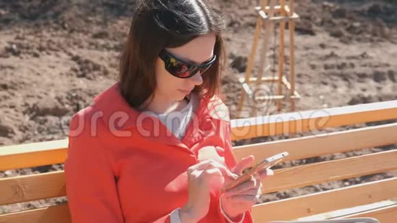 年轻的黑发女孩戴着太阳镜在坐在长凳上的公园里的电话上写信和发送信息视频的预览图