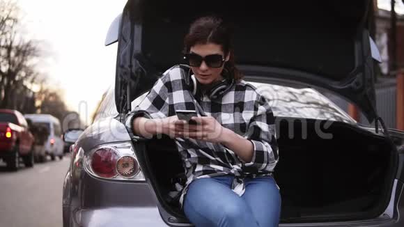 戴墨镜的女人坐在敞开的汽车后备箱里穿着牛仔裤和格子衬衫手机打字视频的预览图
