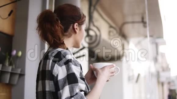 一个穿着格子衬衫的可爱而高兴的女孩站在窗户对面喝着一杯白色的大杯子向外望去视频的预览图