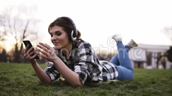 快乐的年轻女性在智能手机上听音乐耳机躺在公园的绿草上女孩利用她的乐趣视频的预览图