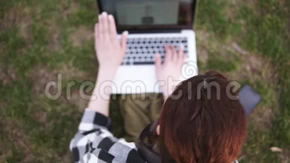 在公园里一个头发尾巴的女孩坐在地上在模糊的笔记本电脑上打印手机在视频的预览图