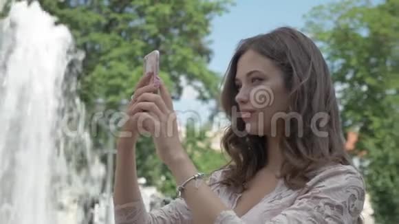 年轻漂亮的长发女孩在电话里拍了一张自己的照片在城市公园喷泉附近视频的预览图
