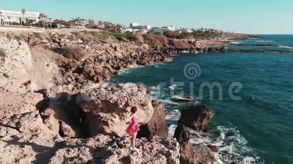 穿着红色衣服的女孩迎风招展双手分开在岩石悬崖上海浪拍打着海岸溅起浪花视频的预览图