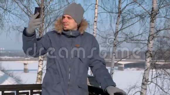 穿着蓝色羽绒服的人带着毛皮罩在冬季公园里用手机进行视频聊天和微笑视频的预览图