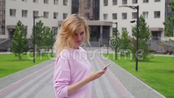 大学背景的年轻女子拿着电话站着穿着粉色毛衣带着手机的女学生视频的预览图