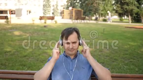 英俊的年轻人带着耳机听着智能手机里的音乐在公园外面的长椅上跳舞视频的预览图