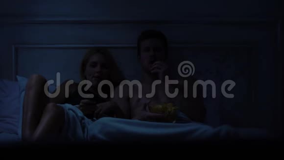 妻子和丈夫坐在床上看电视节目吃零食玩得开心视频的预览图