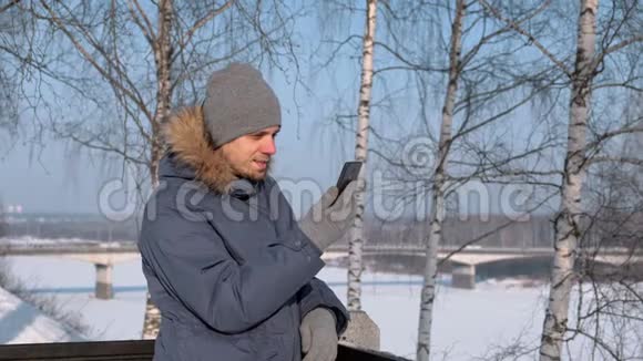 穿着蓝色羽绒服的人带着毛皮罩在冬季公园里用手机进行视频聊天和微笑视频的预览图