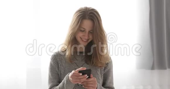 年轻的黑发模特穿着灰色毛衣站在窗户附近的手机上打字视频的预览图
