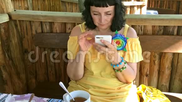穿着黄色衣服的女人在街头咖啡馆的桌子上喝茶用她的智能手机视频的预览图