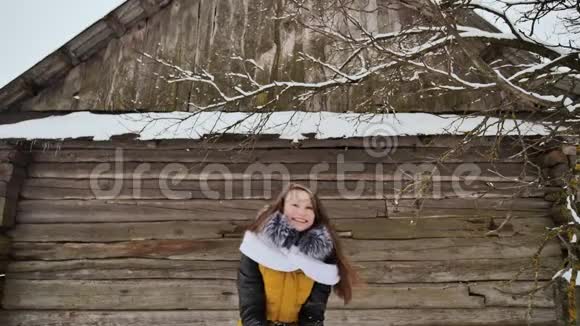 年轻漂亮的女孩玩着扔雪的游戏在冬天的衣服上在木屋的背景上摆着相机视频的预览图