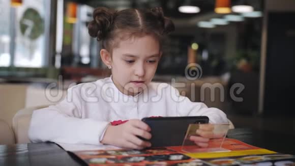 在咖啡馆里一个小女孩热情地在智能手机上玩游戏视频的预览图