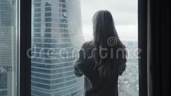 美丽的金发女郎站在豪华的现代公寓或酒店客房的长长的窗户外后视图视频的预览图