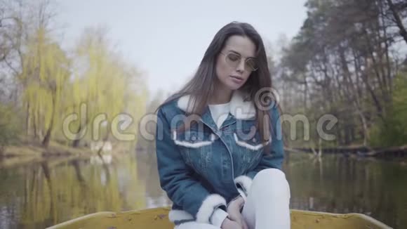 一幅美丽的年轻女子戴着太阳镜和一件牛仔夹克漂浮在湖面或河流上的船上的肖像美丽美丽视频的预览图