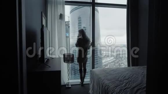 美丽的金发女郎站在豪华的现代公寓或酒店客房的长长的窗户外后视图视频的预览图