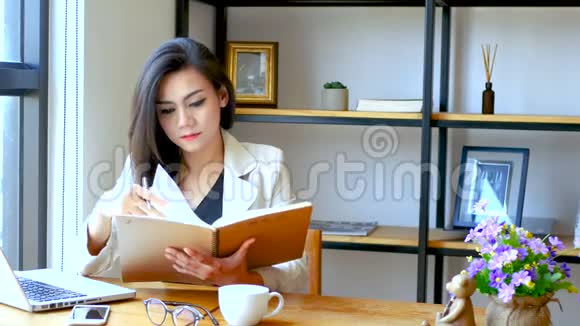4K镜头美丽的亚洲商务女性坐在电脑笔记本电脑前阅读并翻翻笔记簿页面视频的预览图