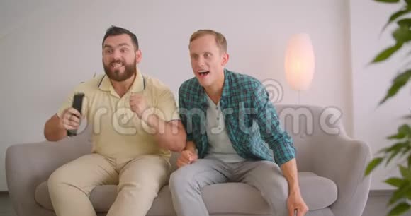 两名白人男子在电视上看足球时坐在家里的沙发上的特写镜头视频的预览图