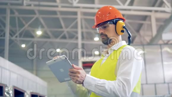 一个戴着安全帽和耳机的工人在他的小工具上键入信息在仓库里看货物视频的预览图