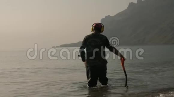一个拿着金属探测器的人正在海滩上寻找有价值的物品黎明时分雾中的悬崖背景下视频的预览图