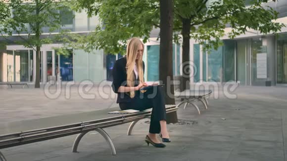 女人在长凳上用平板电脑正式的女商人坐在办公室露台的长凳上休闲地浏览平板电脑视频的预览图