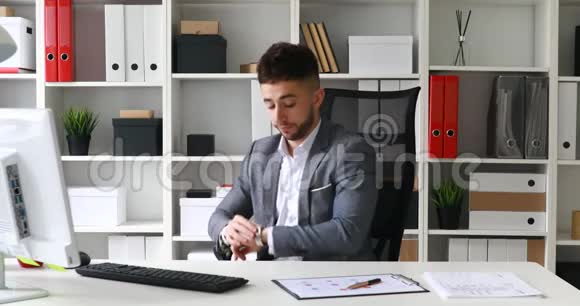穿着灰色夹克的商人坐在白色办公室的桌子旁看着时钟坐在办公室的椅子上离开视频的预览图