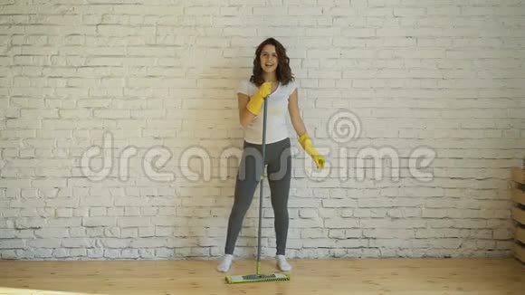 戴着防护手套的漂亮年轻女子正在用拖把唱歌在客厅打扫房间时跳舞视频的预览图