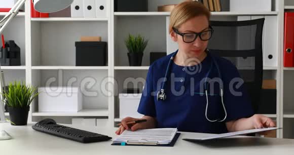 穿蓝色实验室外套的医生坐在工作场所并标记文件视频的预览图